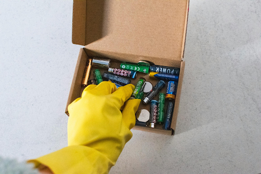 Hand wearing a yellow protective glove placing used lithium-ion and household batteries into a cardboard box for safe disposal, recycling, and compliant hazardous material handling