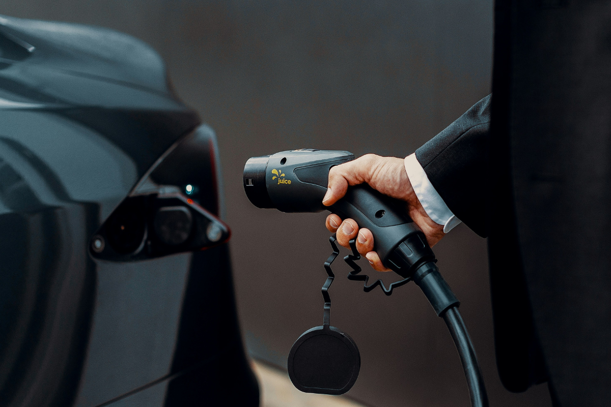 A person in a business suit holds a "Juice"-branded electric vehicle charging connector next to an EV's charging port, representing professional adoption of EV charging technology and infrastructure in a corporate or commercial context.